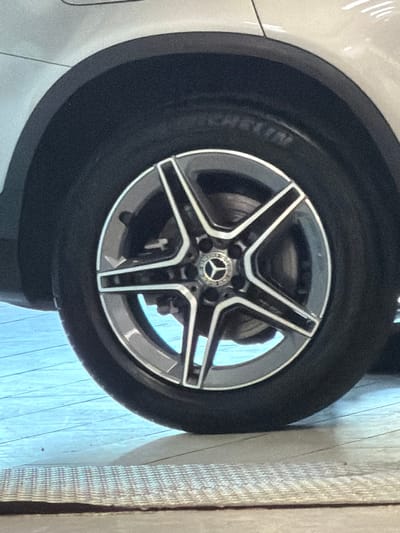The image displays a close-up of the passenger-side wheel of a white or light-colored Mercedes-Benz vehicle, located in Aalsmeer, Netherlands. The wheel consists of a five-spoke, two-tone alloy rim with a Mercedes-Benz star logo centered on the hubcap. The tire is a Michelin brand and appears significantly underinflated or flat, exhibiting a distinct flattening where it contacts the ground surface. A dark, textured fender arch trim is visible above the tire. Through the spokes of the wheel, a brake rotor and caliper assembly are discernible. The vehicle is positioned on a light blue or pale grey floor with visible parallel lines, suggesting a tiled or planked surface. In the bottom foreground, a section of a light-colored, woven mat or carpet runner is present. No individuals or active events are depicted.