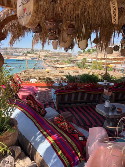 This is an outdoor seating area at a beach resort in Sharm El Sheikh, Egypt. The area is covered by a thatched roof and decorated with traditional Middle Eastern textiles and lanterns. In the foreground, there are several comfortable seating arrangements with plush cushions and colorful rugs. In the background, a beach with sunbathers and umbrellas can be seen, along with the blue waters of the Red Sea and distant hills dotted with buildings. The overall atmosphere is relaxed and inviting, typical of a vacation destination.