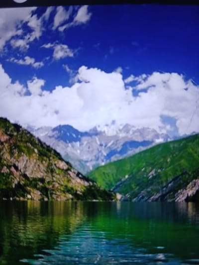 The image depicts a serene mountain lake scene. The foreground is dominated by a body of water with greenish-blue hues, reflecting the surrounding landscape. On either side of the lake are steep, rocky slopes covered in lush green vegetation, indicating a mountainous environment. In the background, majestic snow-capped mountains rise against a vibrant blue sky dotted with fluffy white clouds. The overall impression is one of natural beauty and tranquility. The image does not contain any people or specific architectural elements that would indicate a location within the city of Auk, Kyrgyzstan; it portrays a natural landscape.