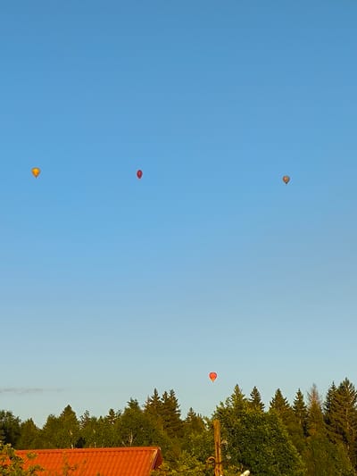 This image depicts a clear blue sky over a dense forest line, with several hot air balloons visible in the distance. In the foreground, the edge of an orange-tiled roof is visible on the left, suggesting a residential area. The balloons are scattered across the sky, with one appearing yellow, two red, and one with striped colors. The overall scene appears to be an outdoor landscape, likely taken during daylight hours. The location is identified as Novonikolskoye, Russia.