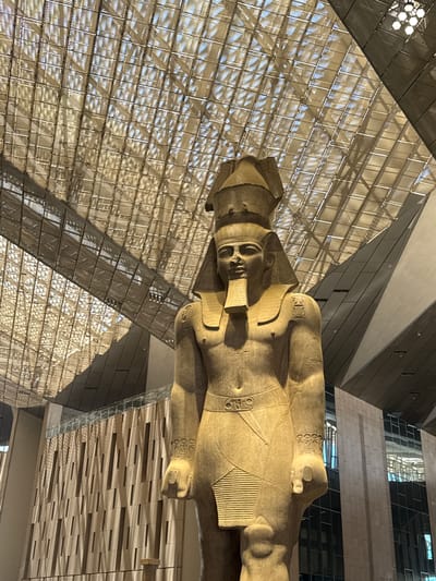 This media file captures a majestic, colossal stone statue of an ancient Egyptian pharaoh, possibly Ramesses II, lying horizontally within a modern architectural space. The statue, carved from light-colored stone, dominates the foreground, with its head, torso, and legs visible. Intricate details are etched into the statue's clothing and headdress, including hieroglyphs and decorative patterns.

The setting appears to be an indoor museum or exhibition hall, characterized by a striking contemporary design. A large, geometric, glass-and-metal ceiling structure creates a grid-like pattern of light and shadow across the scene, suggesting it is daytime with sunlight filtering through. In the background, angular walls and facades constructed from metal panels and glass reflect the light and the surrounding architecture. The overall atmosphere is one of grandeur and historical reverence, juxtaposed with modern design.

The statue is the primary subject, positioned as if it were recently unearthed or placed for display. There are no other people visible in the frame, emphasizing the solitary presence of the ancient artifact. The lighting suggests a bright, clear day, and the location cues, along with the statue itself, strongly point to a site related to ancient Egyptian history, specifically in Kafrat Nassar, Egypt. No visible text is present in the image.