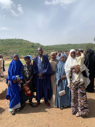 A line of people of varying ages and skin tones stand together in an outdoor setting in Naraguta Hausawa, Nigeria. The line consists of men, women, and children, who are dressed in various forms of traditional Muslim clothing. The women wear hijab and veils of different colors and patterns, including white, blue, beige, and black. The men wear traditional robes and hats, some embroidered. A young child is being held by one of the women. The background consists of a green, sloping hillside on the left and a dirt and grassy ground area. The sky is visible above, with some clouds present.