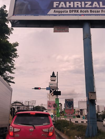 The image presents an urban street scene in Lambaro, Indonesia, from the perspective of a vehicle stopped in traffic. In the foreground, the rear of a red Daihatsu Ayla car is prominently featured, with its brake lights illuminated. To the left, part of a white truck or van is visible, suggesting traffic congestion.
Overhead, a large billboard displays the name "FAHRIZAL" and identifies him as an "Anggota DPRK Aceh Besar," accompanied by social media handles such as "humprodprk_acehbesar" and a website "www.dprkacehbesar.com." Directly below this billboard, a smaller sign advertises "TOWER MEUSEURAYA ADVERTISING" with a contact person and phone number "0853 6273 7207."
In the mid-ground, a red traffic light is visible, confirming the stop in traffic. Below it, a road sign clearly states "DILARANG MENEROBOS" (Do Not Break Through/Do Not Enter). A decorative structure, possibly resembling a minaret or traditional turret, stands near a sign for "BSI Bank Syariah Indonesia." The sky is overcast, providing soft, diffused lighting, indicative of a cloudy day or a specific time like late afternoon.
The background reveals several other commercial billboards and signs along the roadside. Notable among them is one offering "RUKO INI DISEWAKAN" (This shop for rent) with details like "Kanopi depan 15 meter" and a contact number "0812-6933-311." Another sign encourages caution with the phrase "HATI-HATI DI JALAN" (Be careful on the road). The scene depicts a typical Indonesian street environment filled with advertising and traffic infrastructure, momentarily at a standstill.