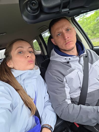 Two individuals, a man and a woman, are seated in the front seats of a car, seen from an upward angle and rotated counter-clockwise. They both appear to be wearing seatbelts.
The man, on the left, has short dark hair, blue eyes, and light stubble on his face. He is wearing a grey and white athletic jacket over a dark blue hoodie. Text on his jacket, which is mirrored, shows "MOBILE" and possibly "TERAVOVA" or similar letters above it. He looks directly at the camera with a neutral expression. Near his head, mirrored text, possibly a VIN or serial number "EB037SSOTTL113", is visible on the car's interior trim.
The woman, on the right, has long brown hair and blue eyes. She is wearing a light blue or lavender jacket and is making a "pouty" or "duck face" expression towards the camera. A black and white patterned bracelet is visible on her wrist, and a blue strap (possibly from a bag) is seen near her.
The interior of the car features dark and light-colored components. Through the car windows, lush green foliage and trees are visible, suggesting an outdoor, natural setting. The reflections on the window and diffused light indicate overcast weather, possibly with some wetness or condensation. An overhead console with a control knob is visible in the car's ceiling. A red seatbelt release button is visible on the passenger side.
The image captures a casual moment of two people in a car, likely taken as a selfie or by someone in the back seat, in Бабстово, Russia.