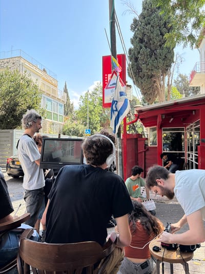 An outdoor film production scene is depicted on a paved street in West Jerusalem, Israel, under a clear blue sky. In the foreground, a person with dark, curly hair is seated with their back to the viewer on a wooden chair, wearing a black t-shirt and white over-ear headphones, monitoring a black video display unit. The monitor shows an image of a clapperboard. To the right of this person, a bearded man in a light t-shirt and glasses adjusts audio recording equipment, including a black sound mixer connected by cables, placed on a small wooden stool. A person with long, dark hair, wearing an orange-brown top, is crouched on the pavement between these two individuals. Further to the right, a man in a green t-shirt is seated near a clapperboard lying on the ground.

To the left of the video monitor, a man with grey, curly hair and a beard, wearing a grey t-shirt and dark jeans, stands and observes the screen. In the background, a red-clad building with large windows, possibly a cafe or restaurant, is visible on the right, with another person partially seen inside. A utility pole prominently stands in the mid-ground, from which an Israeli flag and a smaller rainbow Pride flag hang. A red triangular road sign, indicating a road bump with Hebrew text ("מכשול" and "סיכון") and a blue wheelchair accessibility symbol, is also affixed to the pole. Beyond the red building and utility pole, dense green trees and light-colored stone residential buildings with balconies are visible. A dark sedan is partially visible parked on the street in the far left background.