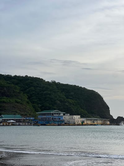 This is a wide shot of a bay in San Juan del Sur, Nicaragua, under an overcast sky. The foreground shows a pebble beach with a small amount of white surf. The mid-ground features a calm bay with several buildings along the shore. The building on the left is light green with a white awning and appears to be a restaurant. To its right is a larger, blue building with "DOCKSIDE" written on it, also appearing to be a restaurant or bar. Further to the right is a light-colored, single-story building with yellow accents, likely part of the port facilities. A few small boats are visible in the water near the dock. The background is dominated by a steep, densely forested green hill that rises dramatically from the water's edge. The overall atmosphere is peaceful and serene.