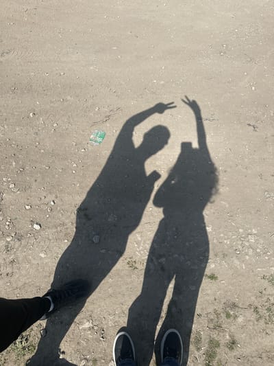 A ground-level view of a section of ground is shown, with the shadows of two people cast upon it. The person on the left is casting a shadow with one arm raised, making a peace sign. There is a small, green and white object lying next to the shadow on the ground. The person on the right is standing close to the edge of the ground, casting a shadow. Their feet are visible with black shoes and white soles. The background is a mix of dirt, small rocks, and vegetation, consistent with a natural outdoor environment. The location appears to be outside of Boryana, Bulgaria.