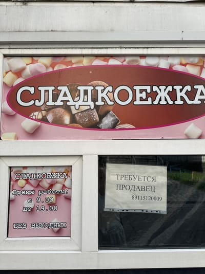 The image shows the storefront of a confectionery shop called "СЛАДКОЕЖКА" (Sladkoezhka). The shop's name is displayed prominently on a large banner with an image of marshmallows. Below the banner, a window displays operational information for the shop. One section reads "Время работы: с 9.00 до 19.00" (Opening hours: from 9.00 to 19.00) and "БЕЗ ВЫХОДНЫХ" (Without days off). Another section of the window displays a sign that says "ТРЕБУЕТСЯ ПРОДАВЕЦ" (Salesperson required) along with a phone number "89115120009". The storefront appears to be located in Rubtsovo, Russia. The overall scene depicts a small retail business with a recruitment notice.