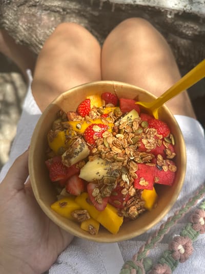 A bowl of fruit salad with granola and seeds sits on a lap. The salad contains watermelon, mango, strawberries, and pineapple. A yellow spoon is stuck into the bowl. The bowl is held by a hand. A flower-patterned strap lies near the bowl.