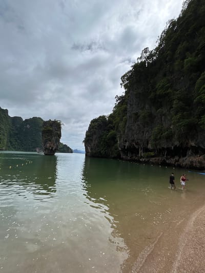 This is a landscape image taken at Krasom, Thailand. The scene depicts a rocky island formation in the middle of the water, with lush green vegetation covering its surface. In the foreground, there is a sandy beach with the water gently lapping at the shore. Two individuals are visible standing in the shallow water near the beach. One person is wearing a red top and white shorts, while the other is in a black t-shirt and dark shorts. The water is a murky green color, and there are some yellow buoys or markers visible in the water. In the background, more rocky islands and the sea stretch out under a cloudy sky. The overall impression is that of a tropical, natural environment. The lighting suggests it might be daytime, possibly overcast given the cloud cover.