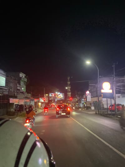 The image depicts a busy street scene at night in Macege, Indonesia. A person's helmet is prominently in the foreground, suggesting the photo was taken from a motorcycle or similar vehicle. The road is illuminated by streetlights and the headlights and taillights of various vehicles, including cars and motorcycles, creating a dynamic and somewhat blurry effect.

The scene is filled with activity, indicated by the presence of multiple vehicles and what appear to be shops or buildings lining the road, many adorned with bright, illuminated signs. A large, circular, orange sign with what looks like a stylized letter "D" or a similar character is visible, possibly a business logo. Red traffic lights are also visible, suggesting the vehicles are stopped or slowing down.

The time of day is clearly night, given the darkness and artificial lighting. The road appears to be wet, reflecting the lights, which could indicate recent rain or humidity. The overall atmosphere is one of a bustling urban environment at night, with the glow of lights and the movement of traffic. There are no explicit indicators of specific emotions, but the scene conveys a sense of a typical, active evening on a Indonesian street.