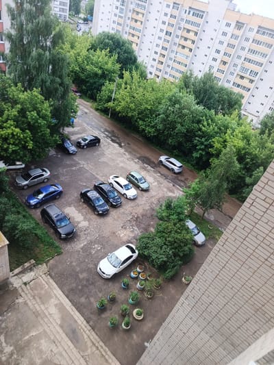 This is an aerial photograph of a residential area in Sof'ino, Russia. The scene depicts a courtyard with several parked cars, a street, and apartment buildings surrounded by lush green trees. The time of day appears to be daytime, possibly overcast given the muted light.
The primary subjects are the cars, which include a white sedan, a dark-colored SUV, a blue sedan, a black sedan, a silver sedan, and a few other dark-colored vehicles. A light-colored vehicle is parked on the far left, partially obscured by grass. There are no people visible in the frame, nor any specific activity taking place, suggesting a still moment in a typical day.
Notable details include the arrangement of the parked cars in designated spots on the asphalt. There are also several decorative planters with greenery scattered around the courtyard, adding a touch of nature to the urban setting. The apartment buildings are multi-story structures with numerous windows, characteristic of Russian residential architecture. The trees are dense and appear to be in full leaf, suggesting a summer or late spring setting. The ground is a mix of asphalt and concrete, with some patches of grass and dirt. The overall impression is one of a quiet, everyday scene in a suburban or urban residential neighborhood. No visible text is present in the image.