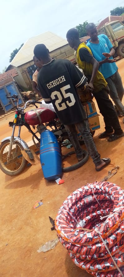 In Baissa, Nigeria, a group of young men are gathered around a red and silver motorcycle parked on a dirt road. The motorcycle has a blue plastic fuel tank attachment. One man in a black t-shirt with "GEHGEH 25" printed on the back stands facing away from the camera, while two others are partially visible. A stack of tires, wrapped in red and silver packaging with "CST" branding, is on the right side of the image. In the background, a beige truck is parked, and various buildings with corrugated metal roofs are visible. The sky is overcast.