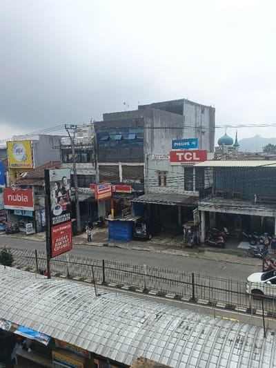 The image depicts a street scene in a somewhat crowded urban environment. Several buildings of varying heights and architectural styles line the street. Visible advertisements include Philips, TCL, WBA, and Nubia. Also, a sign for "PASAR RAKYAT CIPANAS" is seen. Multiple motorcycles and a white car are parked along the street. The sky is overcast, and a mosque's dome is visible in the background. A metal fence runs along the edge of the street, and a corrugated roof is in the foreground.