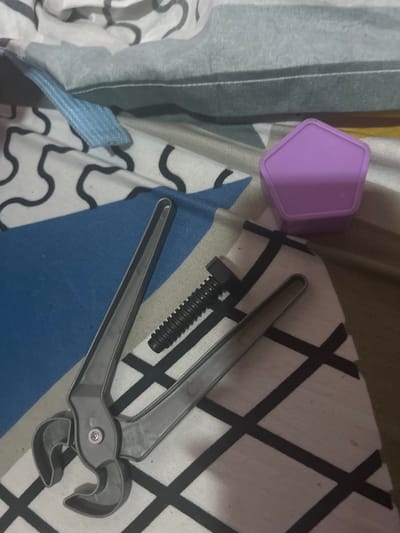 A still life photograph shows a purple pentagonal box, a black screw, and a gray plastic pair of pliers lying on patterned bedding. The background bedding has two different patterns, one featuring white fabric with wavy black lines and the other blue with white crosses. The photograph appears to be taken indoors. The location is unknown and there is no information to confirm that it is in San Juan de Los Morros, Venezuela. There are no people depicted.
