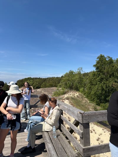 In Morskoye, Russia, on a bright, sunny day, a group of young people are gathered on a wooden boardwalk that overlooks a sandy dune landscape. The boardwalk appears to be part of a nature trail or viewpoint. Several individuals are standing and sitting on the boardwalk, some are looking at their phones, while others are conversing. The vegetation surrounding the dunes is dense with green trees and bushes. The sky is a clear, vibrant blue with a few wispy clouds. The overall scene suggests a leisurely outing in a natural, coastal environment.