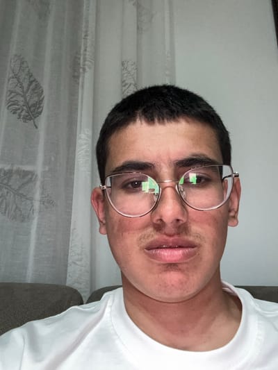 The image displays a single individual, identified as male, located indoors in Kanchevo, Bulgaria. The subject presents with short, dark hair and wears light-colored, possibly gold-toned, framed eyeglasses that exhibit green light reflections. Notable facial features include light stubble on the upper lip and chin, along with areas of redness or blotchiness on the cheeks and forehead. The individual is clad in a white crew-neck t-shirt and maintains a direct, neutral gaze towards the camera. The background consists of a sheer, light-colored curtain adorned with dark leaf patterns on the left side, and a plain, light-colored wall on the right. A portion of a light brown or grey upholstered object is visible at the bottom left of the frame.
