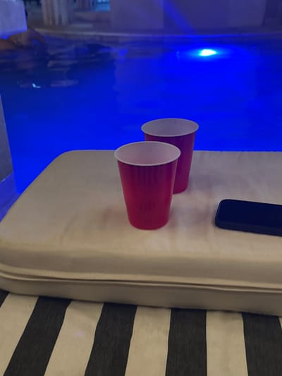 The image shows two red plastic cups and a black smartphone on a white padded surface next to a gray and white striped fabric. To the left of the cups, there is a blue background, possibly a light or reflection. The location, Acapulco de Juárez, Mexico, cannot be determined from the image.