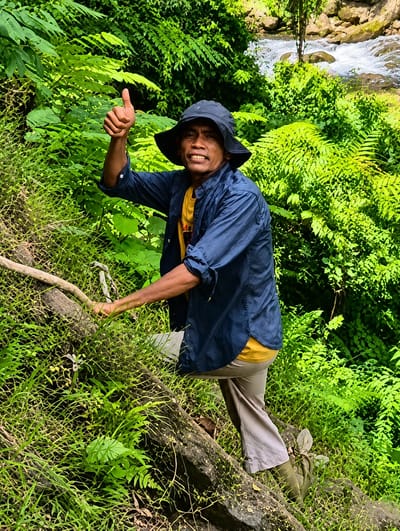 A medium shot captures a man with brown skin and short, dark hair wearing a dark bucket hat, a dark blue button-up shirt over a yellow t-shirt, and light brown pants. He is walking up a grassy incline next to a large, fallen tree trunk. He is looking directly at the camera with a wide smile and his right thumb is raised in a "thumbs up" gesture. The background is filled with lush green foliage and a waterfall with white, rushing water. The scene appears to be outdoors in a tropical environment.