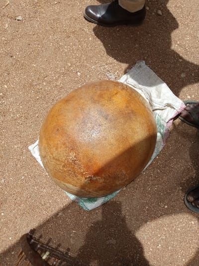 A large, round, brown gourd is resting on a white cloth with green and pink trim. The cloth is on the ground. The ground is covered in small, brown pebbles. A person's brown leather shoe is visible in the upper left corner of the image. The coordinates provided are for the town of Zum, Nigeria. The image appears to have been taken outdoors.