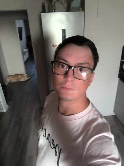 The image shows a man wearing glasses and a light-colored t-shirt, taking a selfie in what appears to be a residential interior. He is positioned in the foreground, with a refrigerator visible behind him to the right. To the left of the frame, a hallway leads to another room where a dog is lying on the floor. The flooring in the visible areas is a dark wood-grain laminate. The overall lighting suggests an indoor environment. The specific location in the city of Kovrov, Russia cannot be determined from the visual information provided.