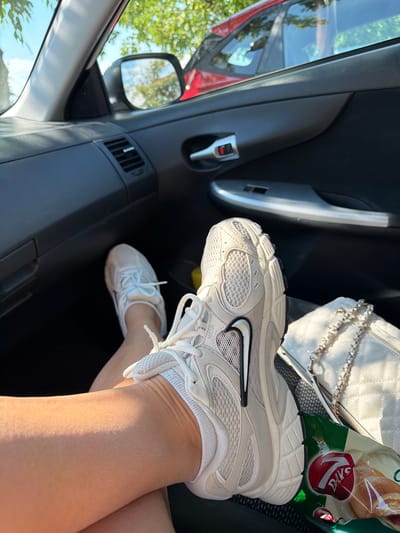 The image shows a close-up view of a person's legs and feet inside a car, with a white quilted handbag and a bag of pastries visible in the background. The person is wearing white athletic shoes with a black Nike swoosh. The car's interior, including the door panel and a side mirror reflecting trees and sunlight, is also visible. The scene suggests a casual outing, possibly a road trip, given the presence of snacks and the setting inside a vehicle. The lighting indicates it is daytime, with bright sunlight filtering into the car, suggesting fair weather. The overall mood is relaxed and everyday. The visible text on the pastry bag reads "7DAYS".
