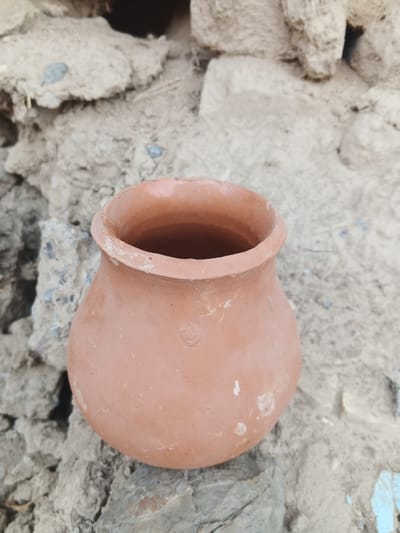 A terracotta pot, appearing to be ancient or rustic in style, rests on a rough, textured surface composed of stones and earth. The background is a wall constructed from similar rough-hewn stones and dried mud, with visible rocks and indeterminate debris. The pot is reddish-brown in color, with a slightly uneven rim and a rounded, bulbous body. A faint circular imprint is visible on the front of the pot. The lighting suggests an outdoor environment, possibly during the day. The image does not contain any people or animals. The location is identified as Farkhad, Uzbekistan, though no specific urban landmarks are visible to confirm this.