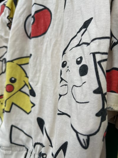 The image displays a close-up view of a white fabric with a repeating pattern of cartoon characters, primarily Pikachus from the Pokémon franchise. The Pikachus are rendered in black outline with yellow fills for some of them, and some have red circular cheeks. The fabric appears to be a piece of clothing, possibly a shirt or a pajama set, due to the visible seams and folds. The background is out of focus and hints at other garments or textures, suggesting the item is likely in a domestic setting or a retail display. There are no people, interactions, or environmental details visible that would allow for identification of a specific location in Haaslava, Estonia.