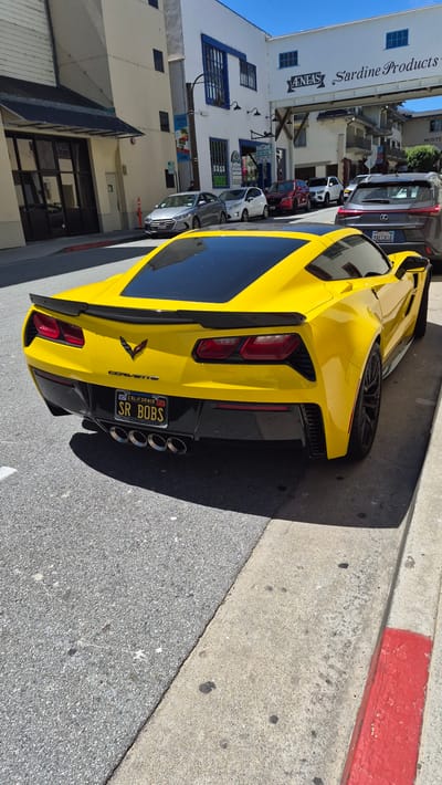 A bright yellow Chevrolet Corvette Stingray is parked on a street in Monterey, California, USA, with its rear facing the camera. The car has a black roof and a black rear diffuser with quad exhaust pipes. Its license plate reads "SR BOBS" and is a California plate. In the background, there are buildings, including one with a sign that says "Sardine Products." Other cars are also parked on the street. The day is sunny, and the scene appears to be a typical streetscape in a coastal town.