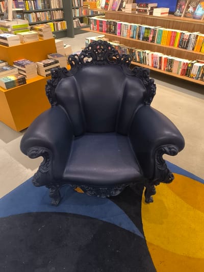 Here is a detailed description of the image:
**Scene:** An ornate, navy blue armchair takes center stage in what appears to be a bookstore. The chair is positioned on a circular rug featuring concentric color blocks of black, blue, and yellow. Bookshelves filled with colorful books line the background, with stacks of books displayed on tiered shelves to the left.
**Location:** The bookstore is located in Leidschendam, Netherlands.
**Objects:**
* **Armchair:** Navy blue, ornately decorated, with a high back and curved armrests.
* **Books:** Numerous books of varying colors and sizes fill the bookshelves and are stacked on shelves.
* **Bookshelves:** Several shelves filled with books.
* **Rug:** Circular rug with concentric color blocks of black, blue, and yellow.
* **Shelves:** Tiered shelves with books stacked on them
**Environment:** An indoor bookstore setting, well-lit with a focus on displaying books in a visually appealing way.