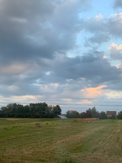 The image displays a rural landscape in Bratkivtsi, Ukraine. The upper portion is dominated by a sky with extensive grey and dark blue cloud cover, interspersed with lighter blue patches and areas exhibiting faint pink and orange hues. Five parallel utility wires traverse horizontally across the mid-ground, positioned approximately one-third down from the top of the frame. Below these wires, a dense treeline composed of dark green deciduous trees extends across the left and central parts of the image.

Beyond the treeline, several structures are visible: a light-colored building with a dark, sloped roof on the left, partially obscured; a building with a reddish-orange tiled roof in the center-right; and a larger building with a dark roof on the far right. The foreground consists of a large expanse of green and yellowish-green agricultural fields, exhibiting patterns consistent with recent mowing or harvesting activity. A small, conical pile of hay is located in the left-center portion of this field. No individuals or animals are present. The lighting is consistent with either dawn or dusk.