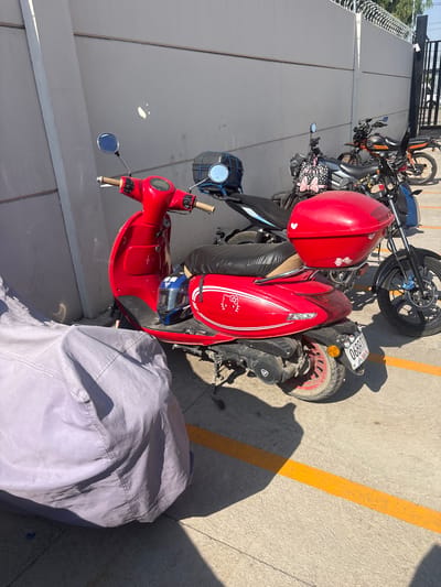 The image captures an outdoor parking area in Buenavista, Mexico, illuminated by bright sunlight, indicating daytime with clear skies. Several motorcycles and scooters are parked on a concrete surface marked with yellow lines. Strong shadows are cast across the ground by the vehicles and potentially nearby structures.
In the foreground, a prominent red scooter, featuring white stripes and a "Hello Kitty" decal on its front panel, is parked. A red helmet with a white bow sticker rests on its seat, and a blue/black helmet is stored in the under-seat compartment. The scooter has chrome mirrors.
Behind the red scooter, a line of other two-wheeled vehicles is visible. This includes a dark-colored motorcycle or scooter with a red seat, another with a black/gray body and a bag decorated with a pink bow pattern near the handlebars, and a motorcycle with an orange fuel tank or accents. Further back, a dirt bike or off-road motorcycle with knobby tires can be seen. A black container secured with blue straps is attached to the handlebars of one of the bikes.
A license plate, "06RRT1," is visible on one of the motorcycles. A stylized logo resembling an 'A' or 'V' within a circle is present on the engine casing of the same vehicle. In the lower right of the frame, a light gray cover or tarp partially conceals an object on the ground. To the left, a wall topped with razor wire suggests a secured boundary. All vehicles appear to be stationary and parked.