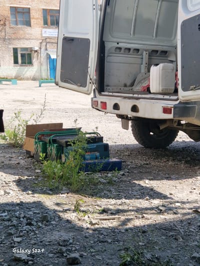 The image depicts an outdoor scene in Zheleznogorsk-Ilimsky, Russia, with a white panel van parked on a gravel surface. The rear doors of the van are open, revealing the empty interior and a white plastic container. In front of the van, a collection of green and black toolboxes, one with a red handle, are stacked on the ground, partially obscured by overgrown grass. A brown cardboard box is also present. In the background, a brick building with multiple windows and a blue door is visible. The overall environment suggests an industrial or service area, possibly a worksite.