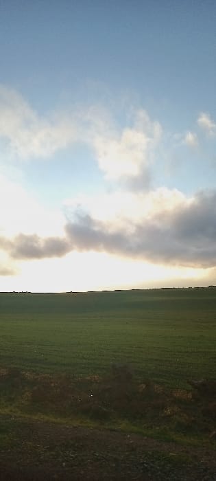 The image displays a rural landscape in Ait Naamane, Morocco, under a partially cloudy sky. The upper two-thirds of the frame feature a light blue sky with scattered white and grey cloud formations. A strong, obscured light source, consistent with the sun low on the horizon, emanates a bright white light horizontally across the middle of the frame, illuminating the undersides of a large, dense cloud bank. A dark, indistinct silhouette line is visible at the horizon, beneath the brightest light, likely representing distant natural or man-made structures. The lower third of the image is dominated by an expansive, uniformly green field, indicative of agricultural land or pasture. The immediate foreground presents a narrow strip of darker, uneven ground, possibly a dirt path or verge, containing minor irregularities and dark patches of sparse vegetation. No individuals, animals, specific objects beyond the landscape, actions, or interactions are visible within the scene. The lighting conditions suggest either dawn or dusk.