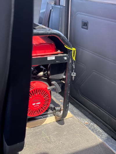 The image shows the interior of a vehicle, likely a utility van or truck, with a portable generator secured inside. The generator is red and black, with the "Fubag GF 420 10.9 hp" branding visible on its red casing. A yellow cord is tied to the generator's frame, and a key is attached to this cord. The generator is resting on a wooden block to stabilize it on the vehicle's floor, which appears to be textured and somewhat worn. The interior of the vehicle shows a dark upholstered panel on the right side, with a recessed handle. The lighting suggests a sunny day, with shadows cast within the vehicle. The general setting suggests a storage or transport context within a rural or suburban area, possibly related to the Мастоцкі сельскі Савет, Belarus, though no specific landmarks or contextual clues point to this precise location within the image itself.