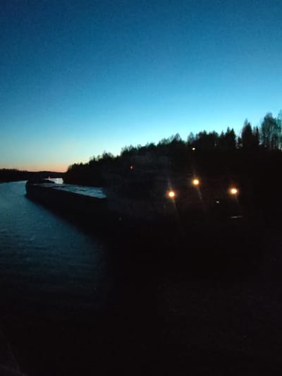 The image depicts a waterfront scene in Uzhla, Russia, at dusk or twilight. The sky is a gradient of blue, fading into orange near the horizon. A body of water, possibly a river, occupies the left side of the frame, with a dark structure or embankment running along its edge. Across the water is a treeline silhouetted against the sky. Further back from the embankment are a few dim lights, with three bright lights illuminating an area to the right of the embankment. The foreground is dark and mostly obscured.