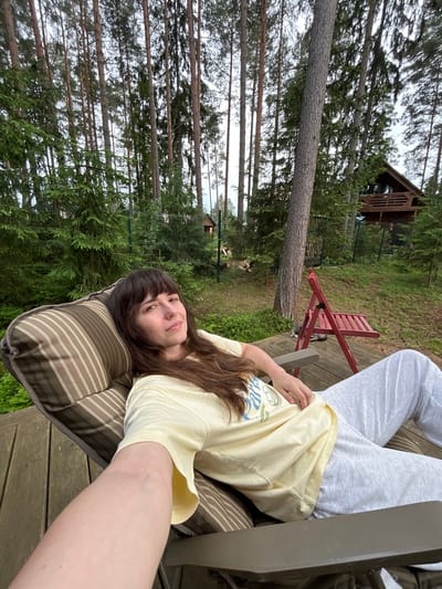 In Shepelyovo, Russia, a woman with long, dark brown hair and bangs is relaxing on a striped cushion chair on a wooden deck. She is wearing a light yellow t-shirt with a graphic design and grey sweatpants. Her right arm is extended forward, resting on the chair. In the background, a dense forest of tall pine trees is visible, with a rustic wooden cabin situated deeper within the woods. A red foldable chair is placed on the deck near the trees, and two cats are lying on the deck in front of it. The overall scene suggests a peaceful, outdoor setting.