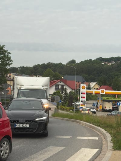 The image depicts a traffic scene on a cloudy day, likely in Limanowa, Poland, given the context of the provided crops and typical Polish road signage. In the foreground, a black Audi sedan with license plate KNS 8222T is stopped at a crosswalk. To its left, the rear of a red car is visible. Behind the Audi, a white delivery truck is partially obscured. Further in the background, a Shell gas station is active, with a white van and other vehicles present. The gas station displays prices of 6.05 and 6.39, likely for fuel. The surrounding environment includes residential buildings with red roofs and lush green trees, indicative of a suburban or town setting. A road sign with the text "KANTON MONEO" is also visible, suggesting a currency exchange service at the gas station. The overall impression is one of normal urban activity, with traffic congestion at a gas station and road intersection.