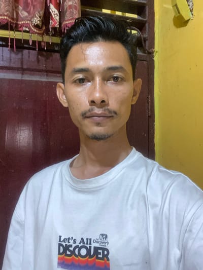 A man with dark hair and a light-skinned complexion poses for a photo indoors. He is wearing a white t-shirt with the words "Let's All Discover" and the Discovery Channel logo printed on it. The background consists of a dark red-brown door or wall, with a yellow wall visible above. To the left, there is a decorative element with red and gold detailing. There is no information available about the location in the city of Tanjongkapai, Indonesia.