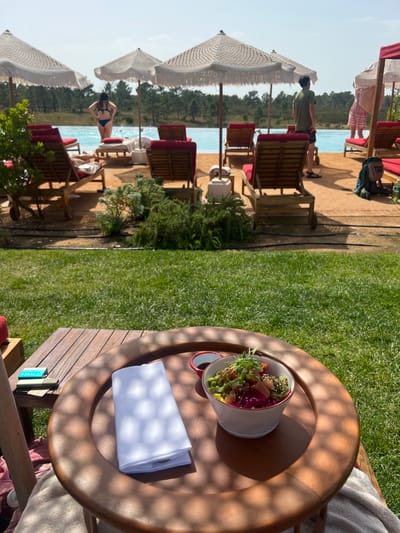 The image depicts an outdoor leisure area at a resort in Aljezur, Portugal, under bright daylight. In the foreground, a round, low-lying wooden table rests on a manicured green lawn, displaying a white folded napkin and a white bowl with a red rim filled with a colorful meal, accompanied by a small dish of dark liquid. A small blue packaged item is also present on an adjacent wooden surface. Mesh-patterned shadows from an overhead structure are cast across the table and lawn.

Beyond the lawn, a narrow strip of green foliage separates the foreground from a light-colored wooden deck. The deck is furnished with multiple wooden lounge chairs featuring red cushions, positioned beneath several white, fringed patio umbrellas. An expansive infinity pool with clear turquoise water occupies the mid-ground.

People are visible on the deck near the pool: one individual in a dark blue bikini stands at the pool's edge, facing away, towards the left. Another individual in a green t-shirt and dark shorts stands further to the right, also facing away from the viewer. A dense forest of evergreen trees forms the distant background, bordering the horizon under a clear, light-blue sky.