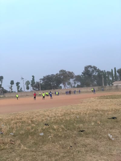 A dirt path stretches along a grassy field. A group of people, some in blue shirts and others in bright yellow vests, are standing on or near the path. Trees and a building are visible in the background. The sky is overcast and hazy. Scattered debris is on the grassy field. The image appears to be taken from a slight elevated angle.