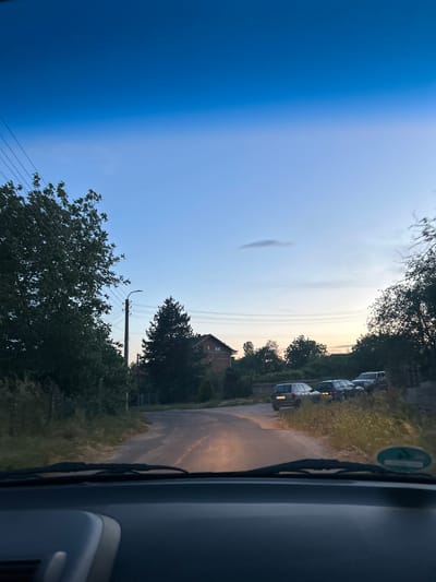The image depicts a view from inside a vehicle, likely a car, driving down a narrow road in Smilets, Bulgaria. The sky is a gradient, transitioning from a deep blue on the left to a lighter, possibly sunset-lit shade on the right. A single cloud is visible in the sky. A row of parked cars lines the right side of the road. A two-story brick building is visible among the trees. Utility poles with power lines are present along the road. Trees and greenery line both sides of the road. The car's windshield wipers are visible.