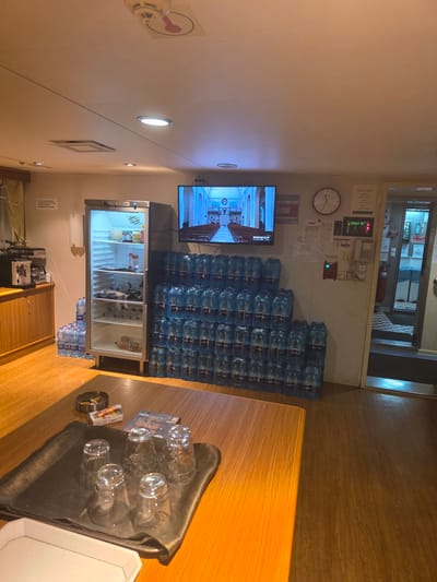 The image depicts the interior of a common area, possibly on a ship or in a facility, given the arrangement of supplies and equipment. The scene is well-lit by overhead lighting and the illuminated television screen.
Dominating the right side of the room is a large stack of blue plastic water bottles, neatly arranged in a pyramid formation. To the left of the water bottles, a tall, stainless-steel refrigerator is open, showcasing its contents, which include various food items and beverages. Next to the refrigerator, a dark wooden cabinet sits below a coffee machine.
A wooden table is positioned in the foreground, on which rests a black tray holding several clear glasses. An ashtray with cigarette butts and a pack of cigarettes are also on the table, along with a video game case.
On the wall, a flat-screen television displays an image of church pews and stained-glass windows, suggesting a religious broadcast. Above the television, a clock is mounted on the wall. A sign with "MEAL TIMES" is also visible. Further down the wall, a red fire alarm and an intercom system are mounted. An open doorway on the right side of the image leads to another area, partially obscured, with a checkered floor pattern.
The environment appears to be a utilitarian space, perhaps a break room or a mess hall. The presence of a large quantity of water bottles suggests a need for hydration, possibly in a location where fresh water is scarce or for a group of people. The overall impression is one of practicality and provision. There are no people visible in the image.
The location is not the city of Brindisi, Italy.