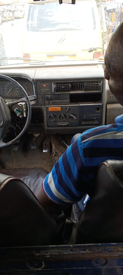 The image captures an interior view from a vehicle, looking out through the windshield at the road ahead. The primary subject is a person, likely the driver, whose right shoulder and arm are visible in the foreground, wearing a blue and white striped polo shirt. The person's head is turned slightly to the right, obscured from full view.
In the mid-ground, the vehicle's dashboard and steering wheel are prominent. The dashboard appears utilitarian and worn, featuring a speedometer and other gauges on the left, an empty radio slot, air vents, and various control knobs and buttons for heating/ventilation. Exposed wires are visible near the steering column, suggesting modifications or wear. The foot pedals are visible on the floor below the dashboard.
Outside the windshield, a bright yellow van or bus is directly in front, partially obscured by reflections. This vehicle has "LAGOS" visible on its rear, likely part of a license plate or vehicle branding, and "Mudo" is partially visible below. In the far background, blurred figures and other vehicles suggest a bustling street or urban environment.
The scene depicts a daytime setting, indicated by the bright ambient light. The weather appears clear. The overall activity is a moment from inside a vehicle, possibly in traffic or waiting, in an urban area. Given the "LAGOS" text on the vehicle ahead and the provided context of "Alasia, Nigeria," the scene is situated in a Nigerian city. Visible text includes numerical readings on the dashboard gauges (e.g., "005480" on the odometer) and the aforementioned "LAGOS" and "Mudo" on the vehicle in front. The interior condition suggests a working vehicle rather than a private passenger car.