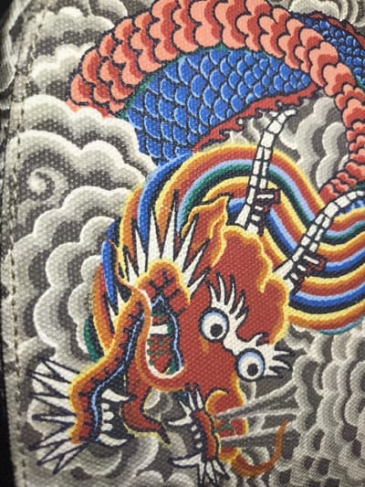 The image presents a close-up, high-resolution view of a printed textile featuring a stylized East Asian dragon motif. The dragon occupies the central and right portions of the frame, rendered in vibrant colors including red, blue, orange, yellow, green, and white. Its head is predominantly red with white, spiky protrusions, prominent round eyes with blue sclera and black pupils, and a flame-like aura in orange and yellow. A multi-colored arc, resembling a rainbow, emanates from the dragon's mouth area. Segments of its body display blue and red scales, and white, segmented limbs with black outlines are visible. The background consists of stylized grey and white cloud or wave patterns. The visible texture of the image indicates a woven fabric surface, suggesting a printed canvas or similar material. A dark vertical seam or edge is present along the left side of the frame. No individuals, specific actions, or environmental cues that would establish a physical location, such as the city of Liège, Belgium, are depicted within the image; the scene is strictly an illustrative pattern on a textile object.