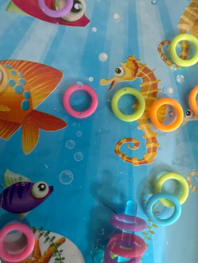 This media file shows a close-up view of a blue surface with a playful underwater theme, featuring cartoon drawings of seahorses and fish. Scattered across the surface are several brightly colored plastic rings. These rings, in shades of yellow, orange, light green, pink, and purple, appear to be toy rings, possibly part of a game or craft. Some are individual, while others are linked together or placed near the cartoon sea creatures. The lighting suggests it is daytime, and the setting appears to be indoors, possibly a play area or a craft table. There is no visible text in the image. The overall impression is one of fun and playfulness, with the vibrant colors and cartoon imagery creating a cheerful atmosphere. The context of Kārsava, Latvia, provides a geographical location for this scene.