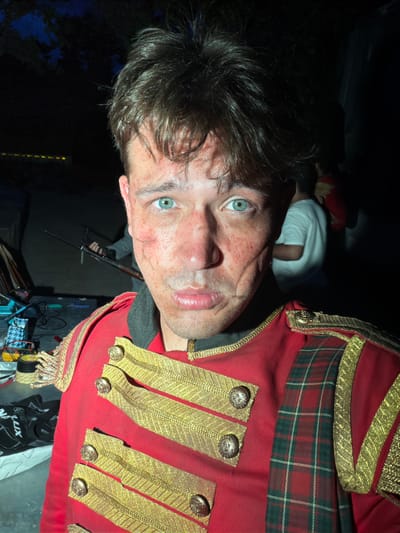 A man with striking blue eyes and apparent face injuries, possibly theatrical makeup, poses for a selfie outdoors at night. He is wearing a red military-style jacket adorned with gold braiding and buttons, and a plaid fabric insert. In the background, other individuals are visible, and there are some objects that suggest a backstage or production area, including what appears to be equipment and props. The scene is dimly lit, with some artificial lights in the distance. The location is stated to be Gollagutta, India, though no specific landmarks within Gollagutta are identifiable from the image.