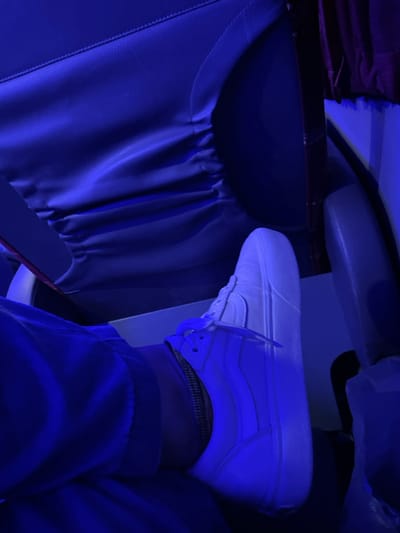 A white Vans sneaker rests on an airplane tray table, positioned close to the seat in front. The seat is covered in a shiny, dark blue material with gathering along the top edge. The blue hue dominates the scene, obscuring details. Part of a person's leg is visible, wearing jeans and an anklet, suggesting someone is relaxing in their seat on the plane.