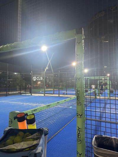 The image displays an outdoor padel tennis facility at night, identified as RGV Padel Club in McAllen, Texas, USA. The entire scene is brightly illuminated by multiple overhead floodlights.
In the immediate foreground, a grey portable cart is filled with numerous yellow padel balls. Two brightly colored ball retrieval tubes, one orange and one yellow, are positioned upright on the cart. A black padel net with a green top band, branded "RGV Padel Club," extends across the lower midground. A vertical metal support pillar, covered in green protective padding also featuring the "RGV Padel Club" logo, is prominent on the right side, partially obscuring a black trash bin.
The padel courts feature blue playing surfaces enclosed by dark wire mesh fencing and glass panels. Two adult males are standing on an adjacent padel court further back, positioned near the net. One individual wears a white top and dark shorts, while the other wears a dark top and dark shorts. Their attire is consistent with athletic activity. In the background, a banner advertising "CLS Commercial" with the slogan "WORLDWIDE DELIVERY" is visible on the perimeter fence. Another illuminated structure in the distant background displays the text "BULLDOGS." Palm trees and ambient city lights are discernible under the dark night sky.