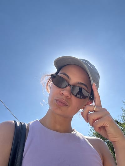 A young woman is captured in a low-angle, close-up selfie outdoors, with a bright blue sky dominating the background. She is wearing a light purple tank top, a grey baseball cap, and dark, oval-shaped sunglasses. The sun is positioned behind her head, creating a halo effect and making the details of her face and hat slightly obscured. Her right hand is raised, with her index finger touching the brim of her cap, and she is wearing a ring with several small stones on her ring finger. Her expression is a slight pout. A black bag is visible on her left shoulder. The scene appears to be a sunny day. The image does not provide any explicit visual information to place it in Lajatico, Italy.